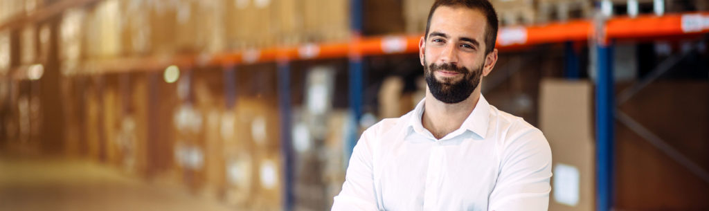 A man stands in a warehouse with his arms crossed and laughs into the camera