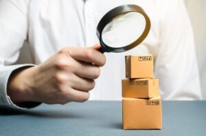 Exporting across EEA borders. A man holds a magnifying glass above the boxes. Examination of goods for the presence of contraband, prohibited goods, defects. Quality control, authentication. study of consumer sentiment