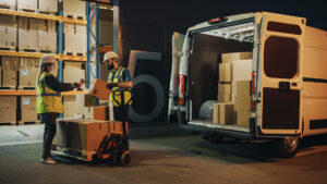 Outside of Logistics Retail Warehouse: Manager Using Tablet Computer and Scanner, talking to Worker Loading Delivery Truck with Cardboard Boxes, Online Orders, Medicine Supply, E-Commerce. Evening